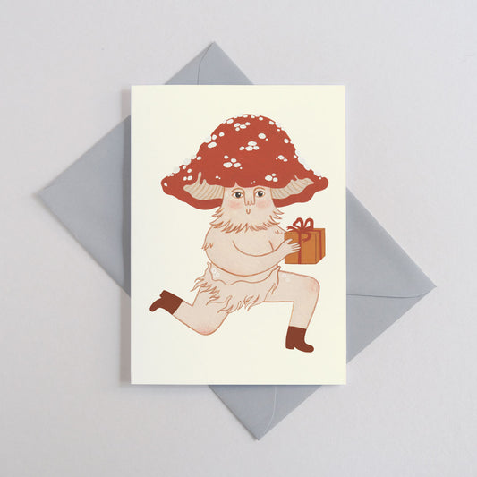 Greeting card featuring a smiling mushroom person holding a wrapped gift. On a grey envelope.